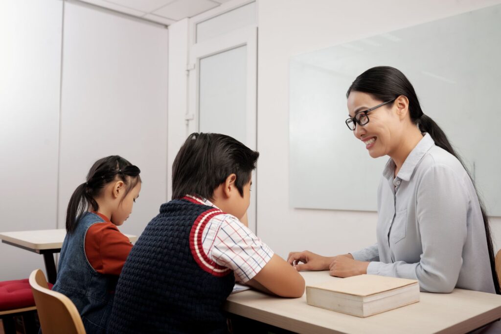 Two asian kids sitting classroom smiling teacher glasses talking boy