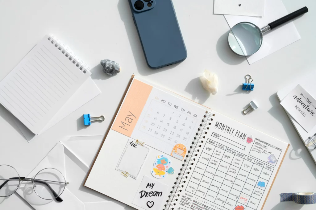 俯拍桌面上的月曆手帳與行事曆規劃頁面，周圍擺放手機、放大鏡與文具，呈現時間區塊安排情境